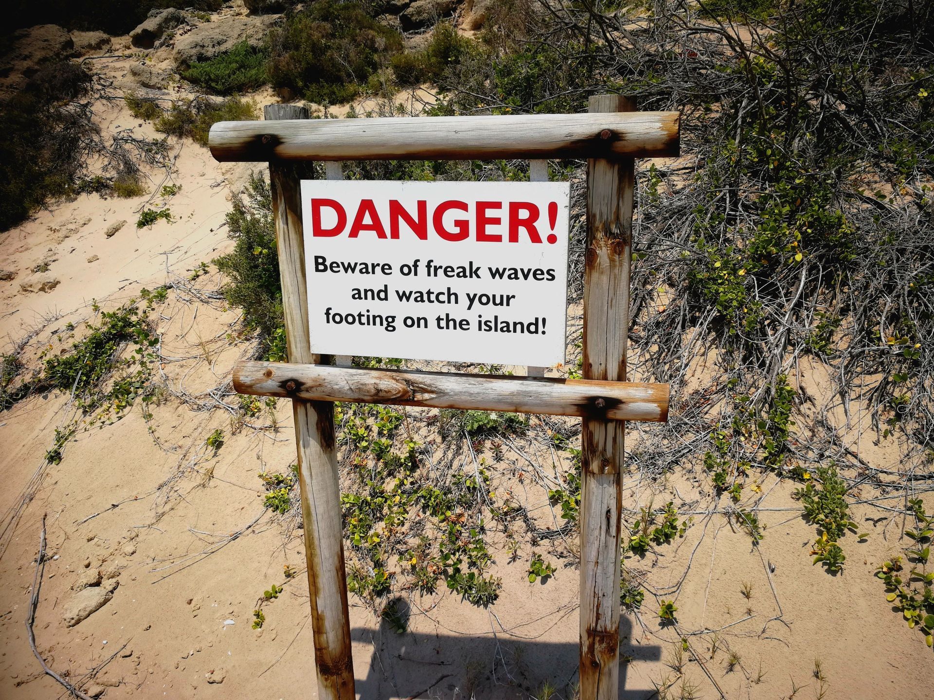 Warning sign on a sandy path: “DANGER! Beware of freak waves washing over your feet!”