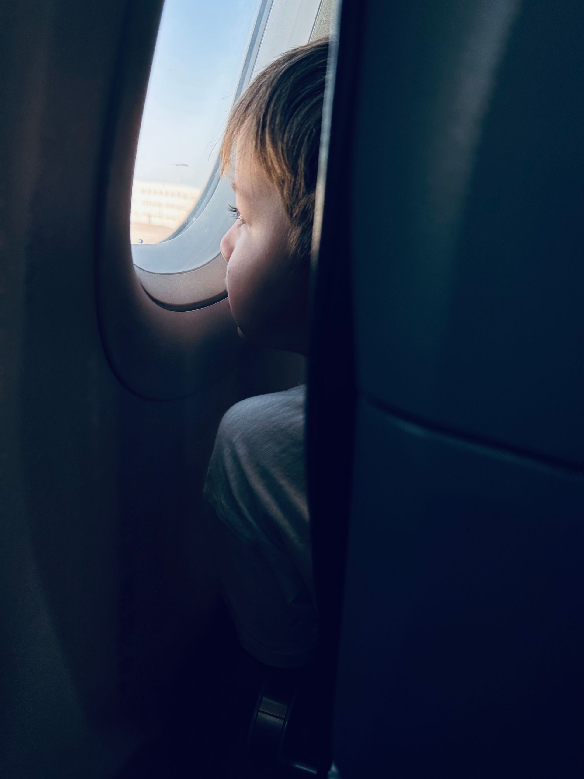 Child looking out an airplane window during daylight, with clouds visible outside.