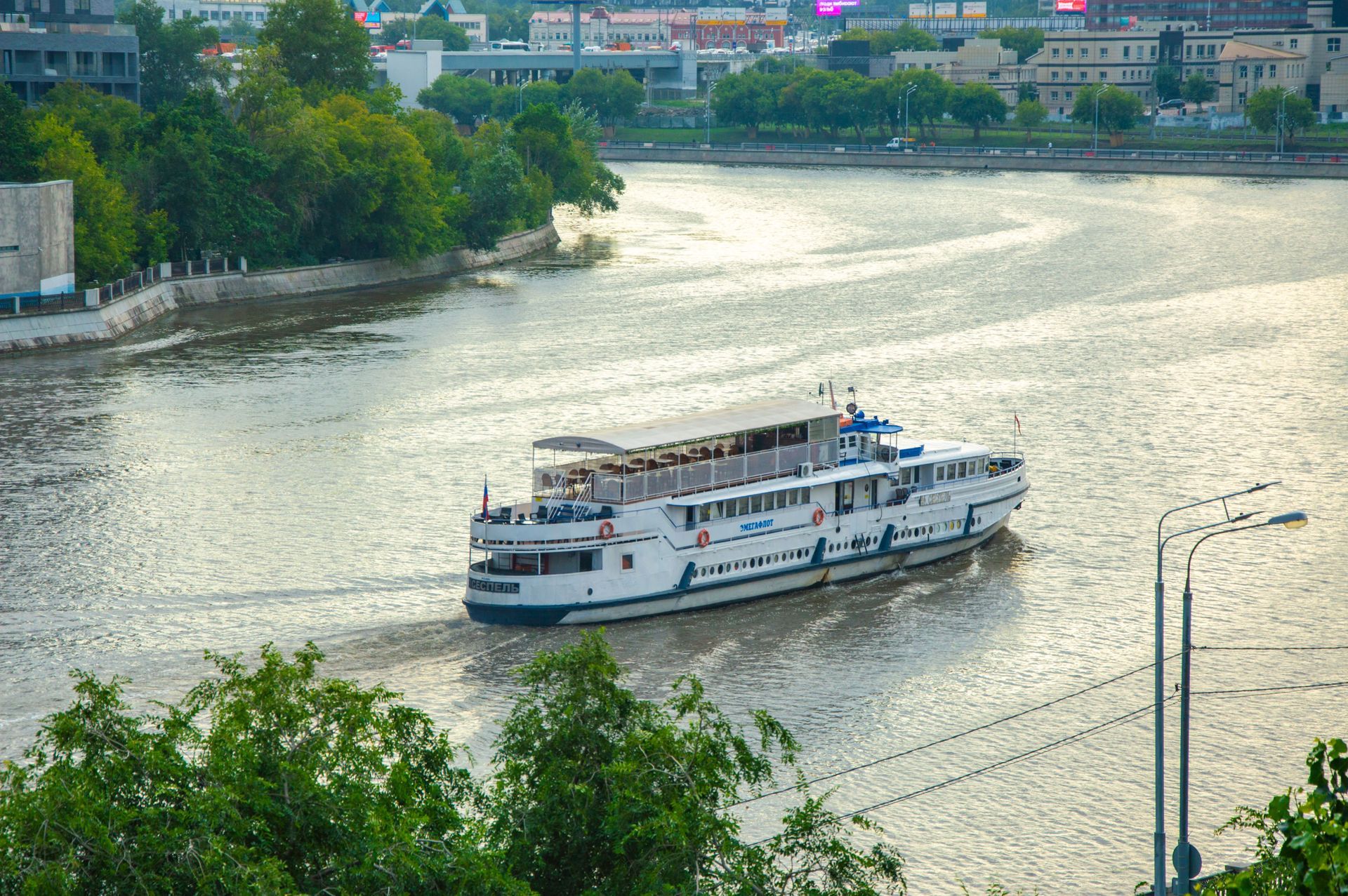 White riverboat cruising on a wide river with green banks and city buildings in the background