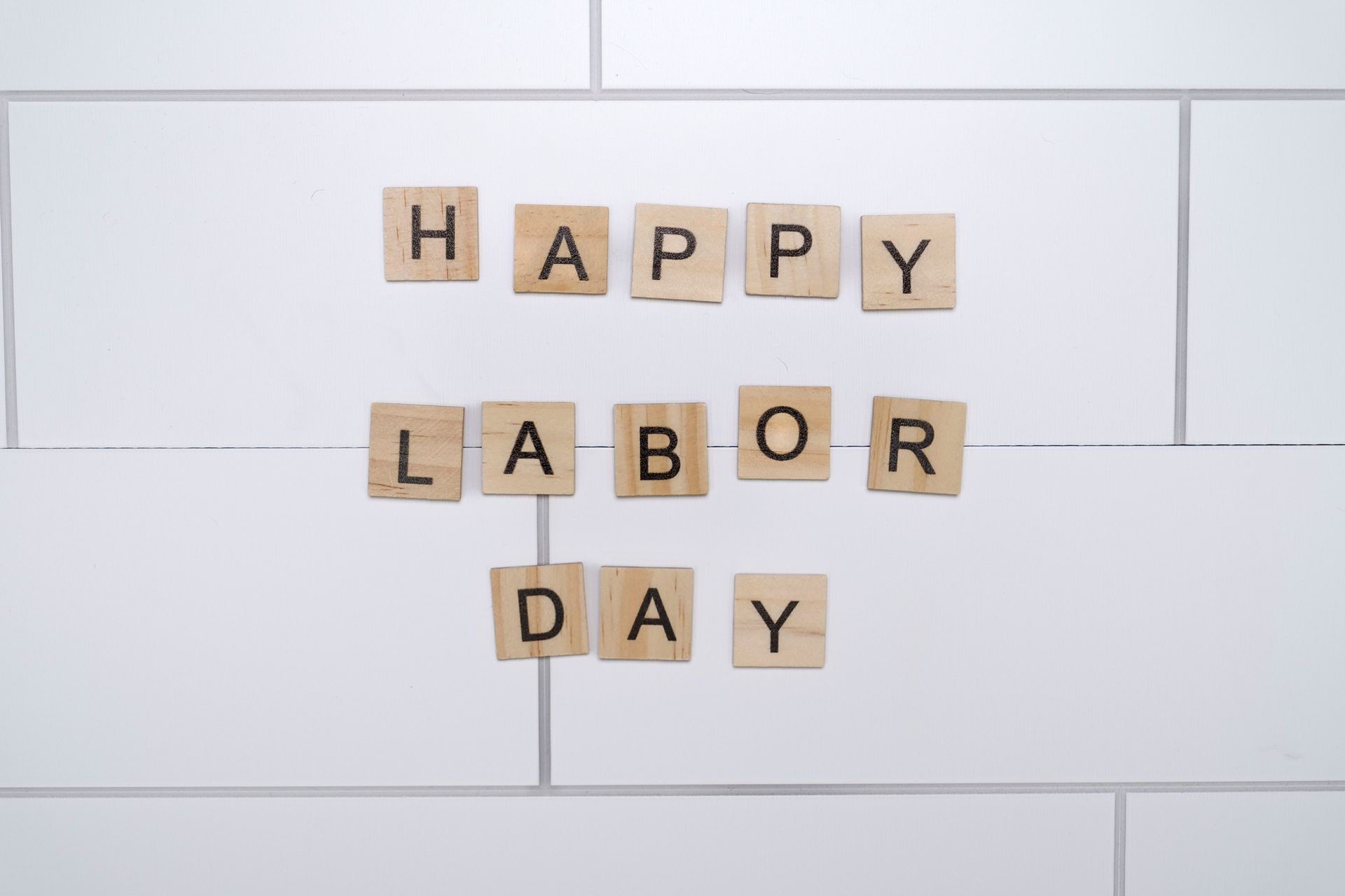 Wooden letter tiles spell “HAPPY LABOR DAY” on a white tiled background.