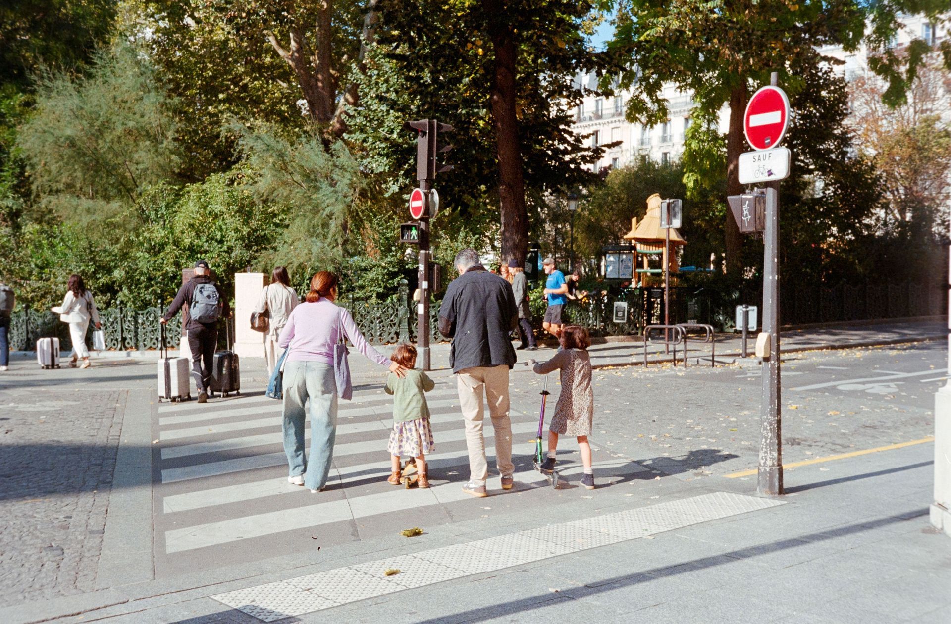 People crossing a street at a crosswalk in a tree-lined urban area, with road signs nearby.