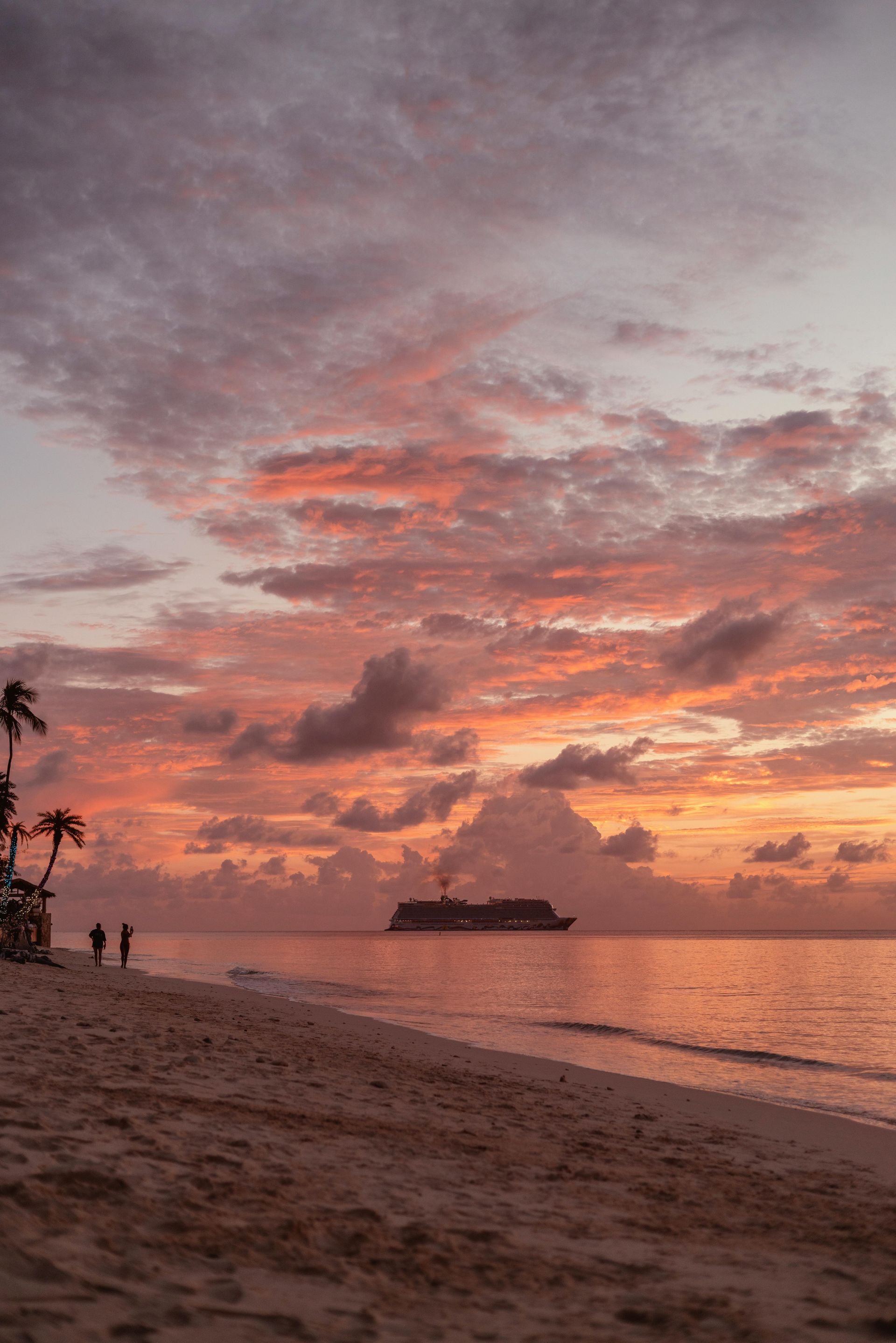 A large cruise ship anchored in the ocean under a vibrant orange and purple sunset, viewed from a sandy tropical beach.