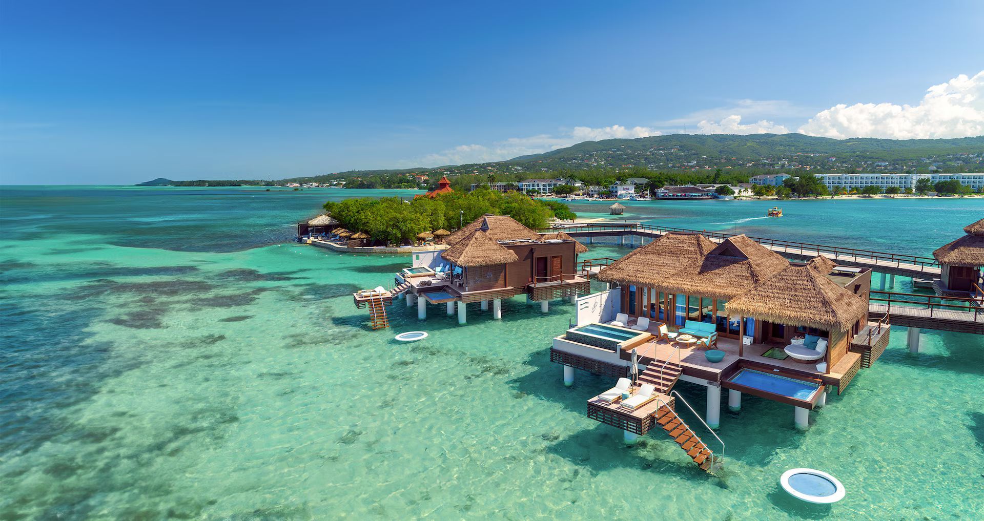 Overwater bungalows with thatched roofs situated in clear, turquoise tropical waters near a coastal island resort.
