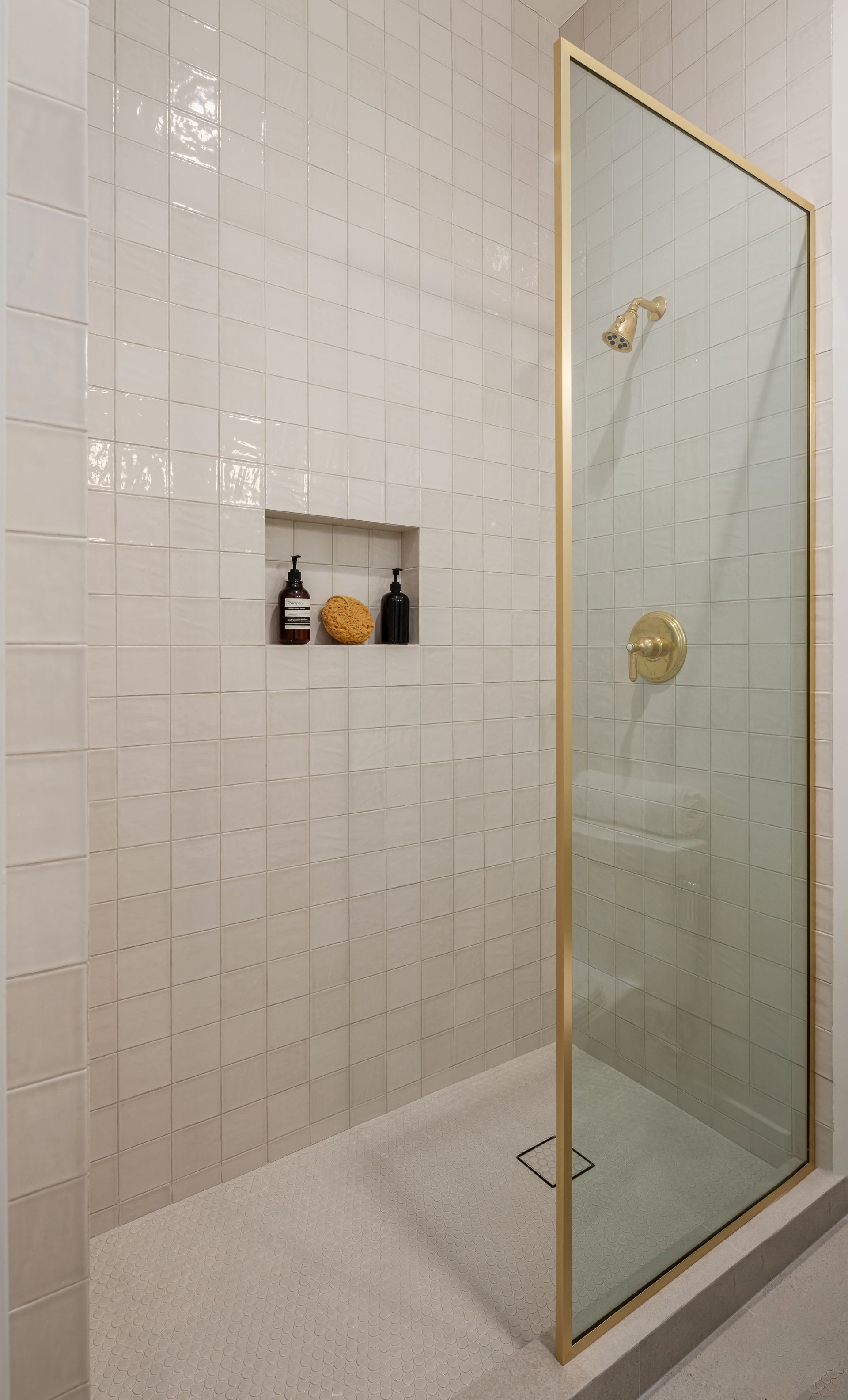 A bathroom with white tiles and a walk in shower with a gold frame.
