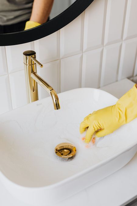 Gloved hand cleaning a white bathroom sink with a gold faucet and drain cover