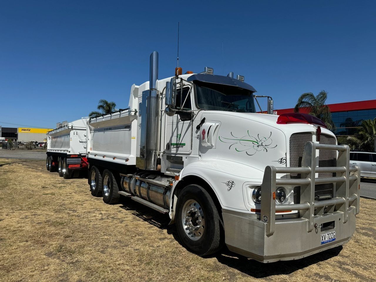 A white Kenworth dump truck with a trailer parked on a dry, grassy lot under a clear blue sky.