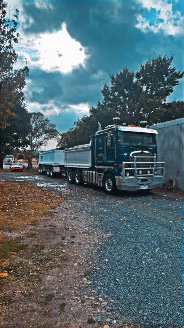 A dark blue Kenworth semi-truck with a silver bull bar and a dump trailer parked on a gravel lot under a cloudy sky.