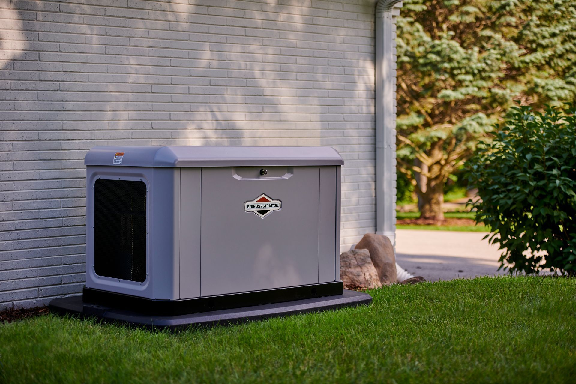 Gray home generator beside a house on a green lawn, with trees and shrubs nearby.