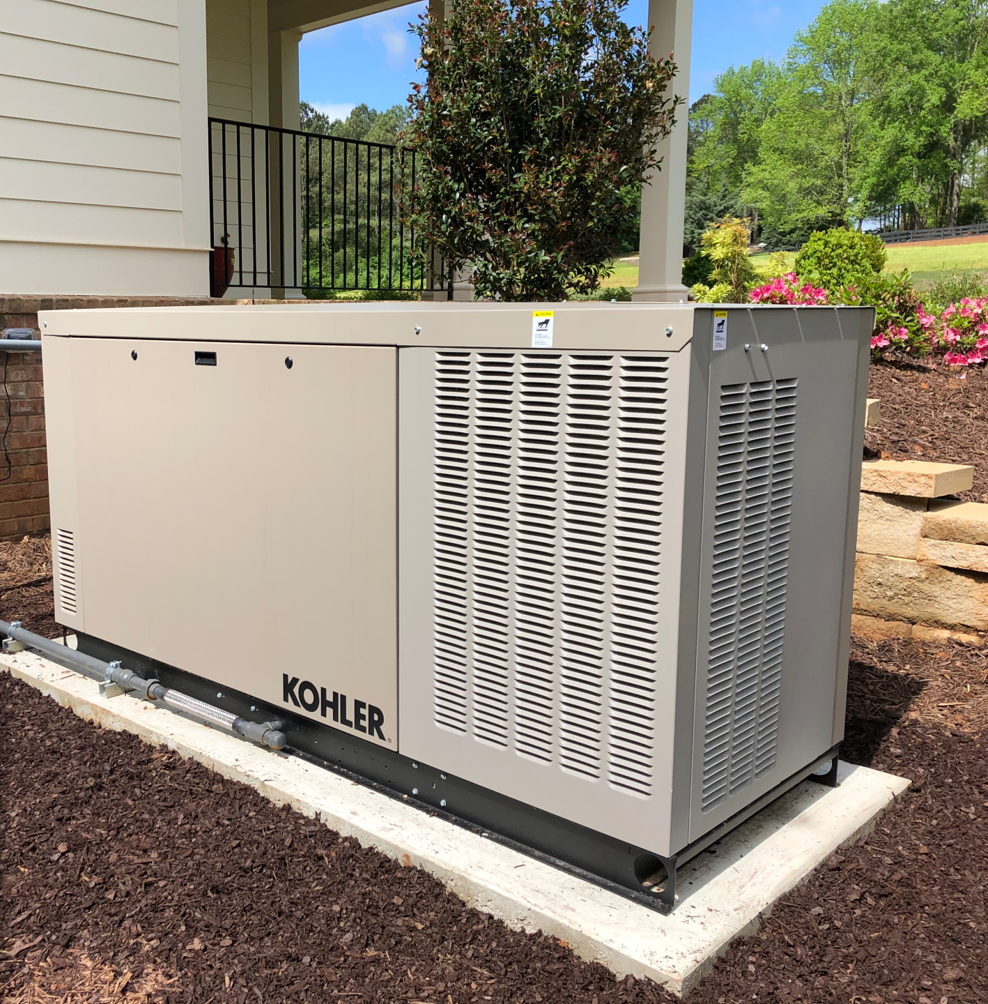 A beige Kohler generator on a concrete pad near a house and landscaping.