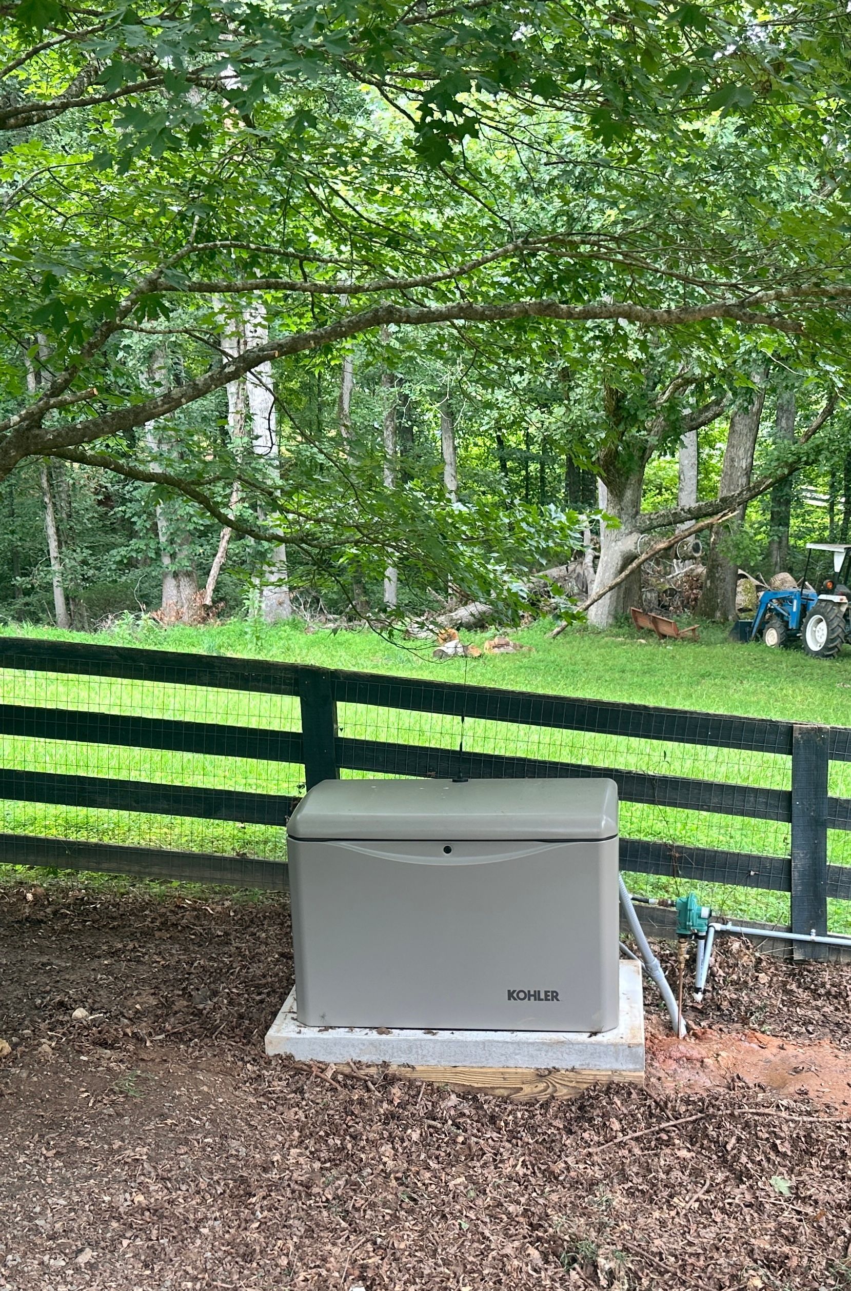 Gray generator on a concrete pad near a black fence, beneath a tree.