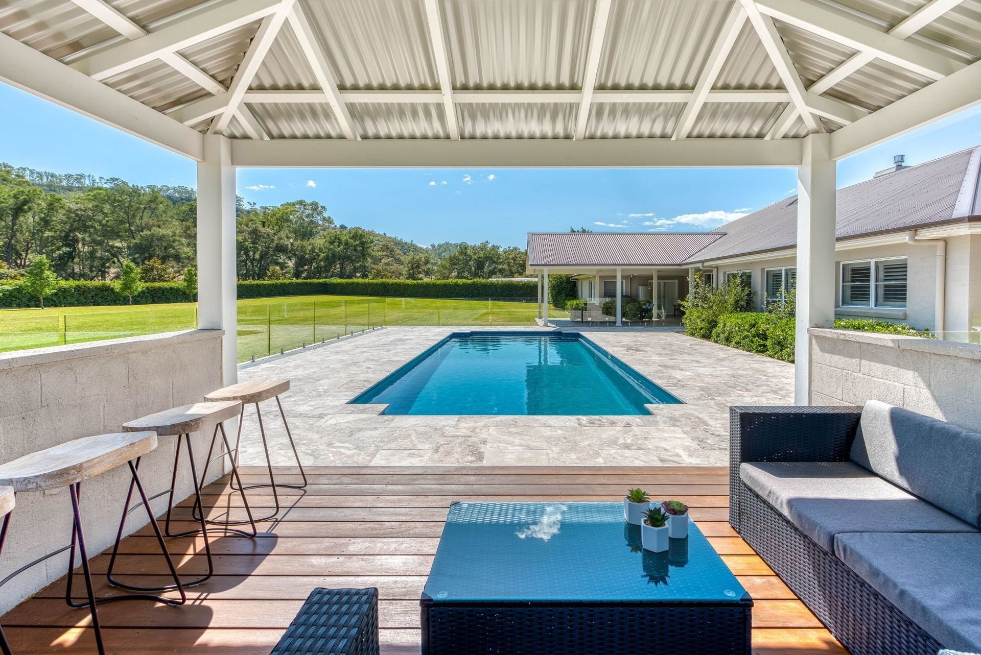 Patio with pool view: pergola, outdoor seating, blue pool, lush greenery, and sunny sky.