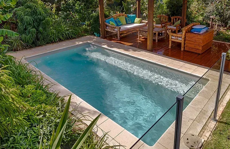 Rectangular pool next to a wooden deck with seating under a pergola, surrounded by greenery.