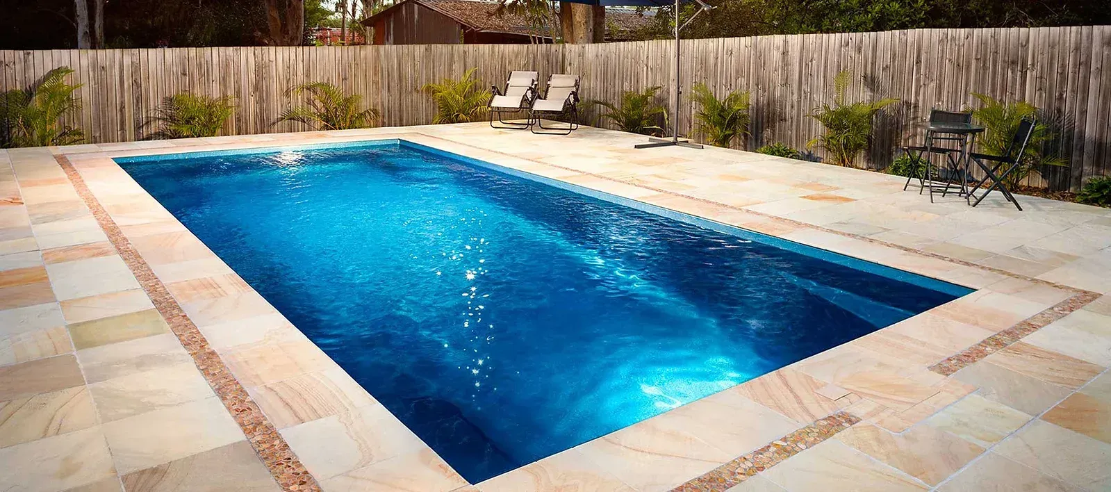 Rectangular swimming pool with blue water surrounded by light-colored stone and a wooden fence.