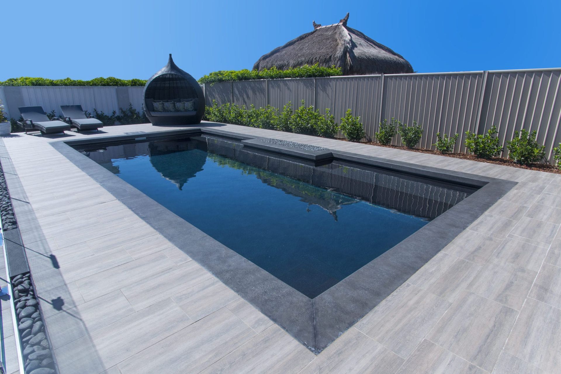Rectangular pool with dark water, light wood-look patio, lounge chairs, and a thatched roof structure in the background.