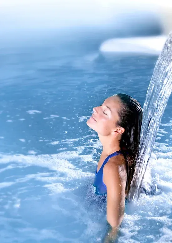 Woman enjoys waterfall in a blue spa, eyes closed with a smile, water cascading over her.