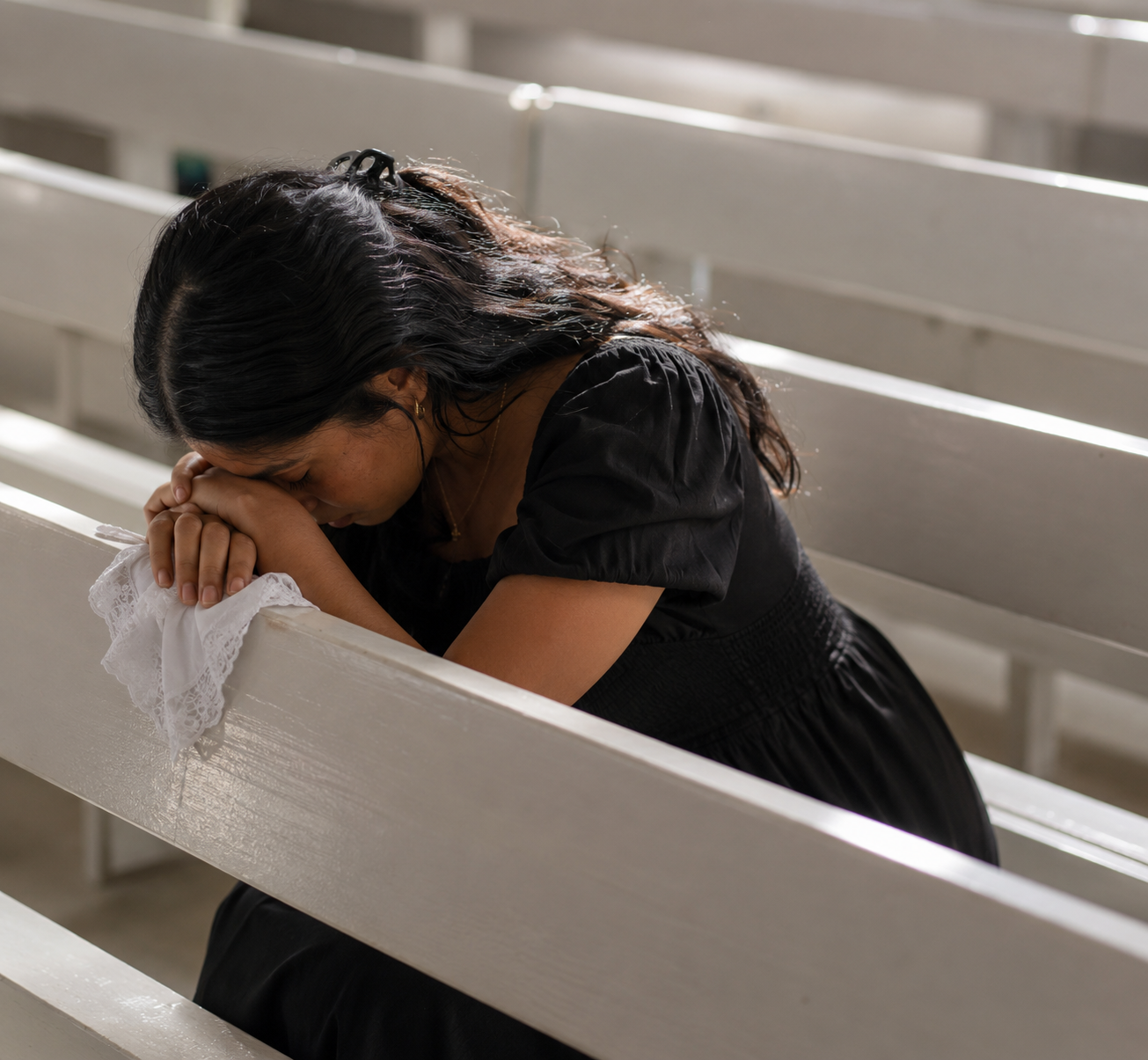 Woman in a black dress kneels in a church pew, head bowed and hands clasped in prayer.