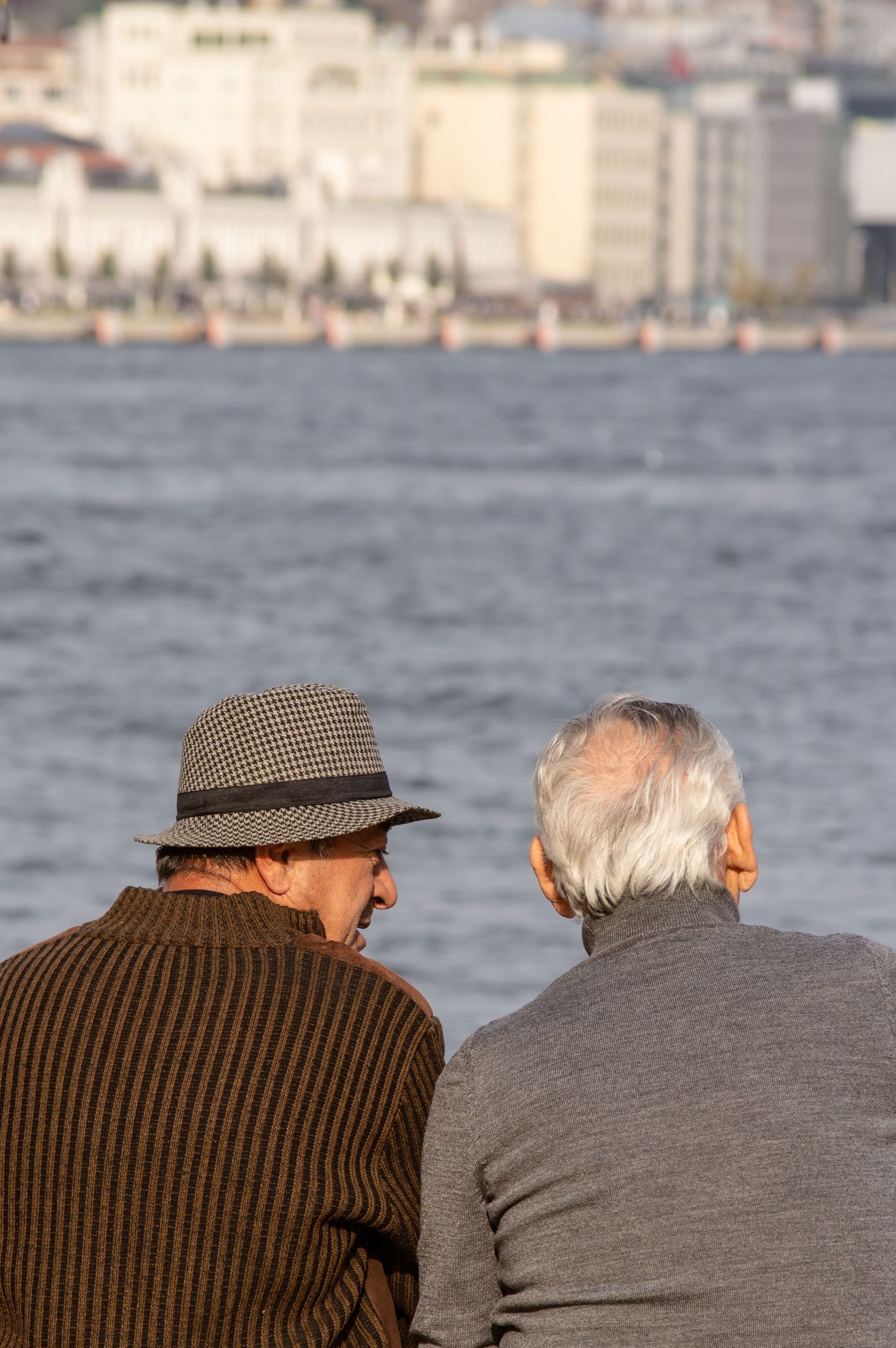Two people sit by the water, seen from behind, with a city skyline in the background.