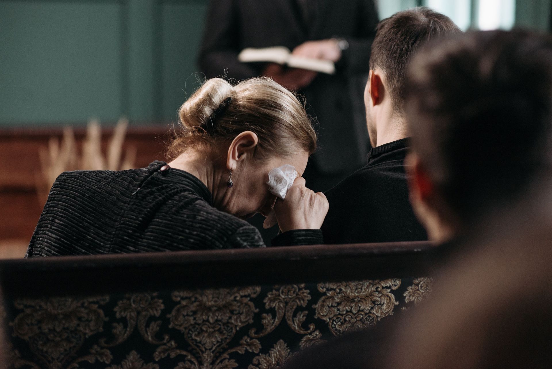 Mourner in black wipes tears in a dim church or chapel, seen from behind pews