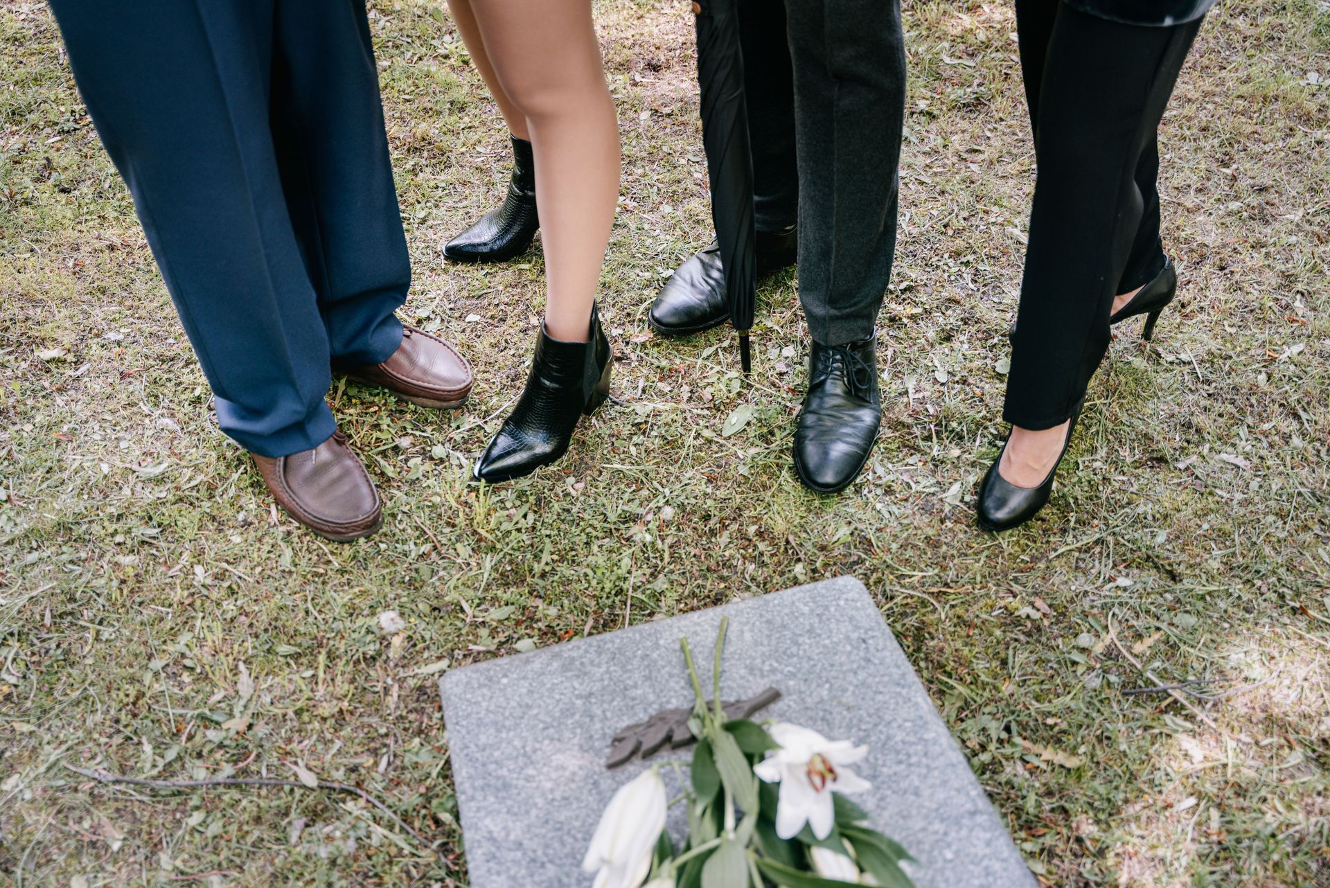 Four people’s legs and feet standing around a grave marker with flowers on grass