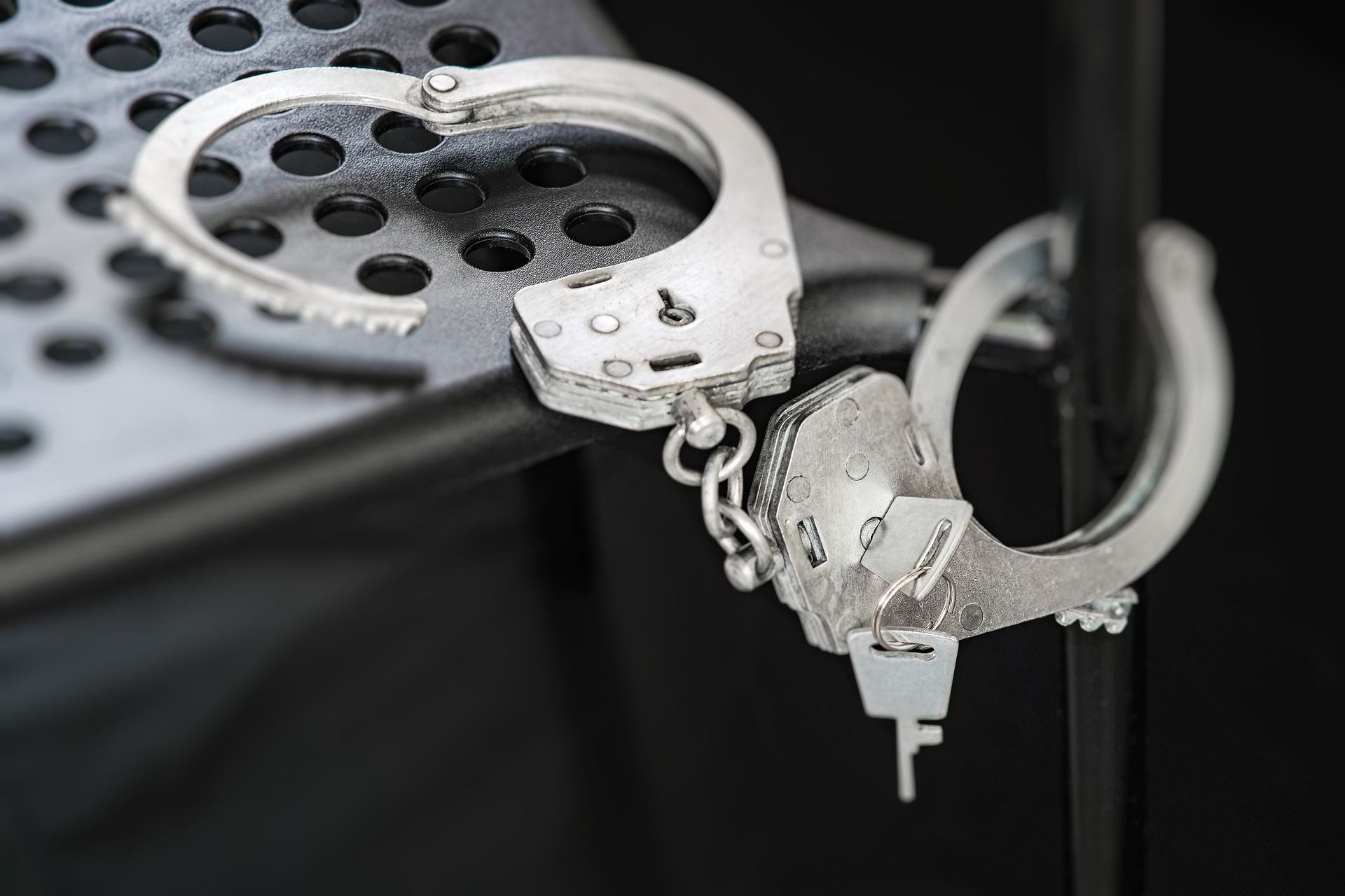 Silver handcuffs resting on a perforated metal surface, with one cuff open and a key attached.