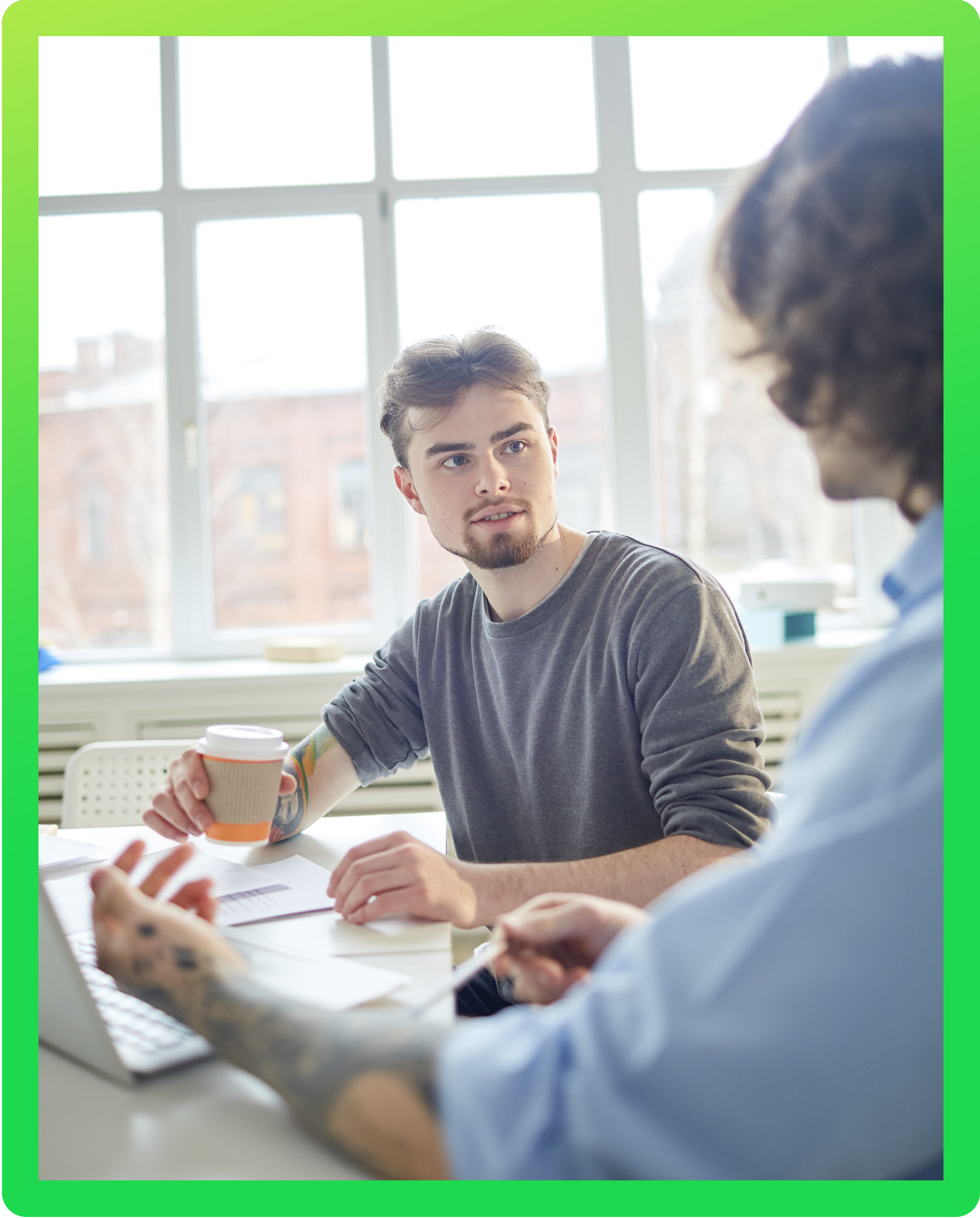 Dois homens estão sentados a uma mesa com um laptop e uma xícara de café.