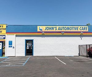 John's Automotive Care storefront with blue, yellow, and red signage. White building with blue parking lines and a sunny sky.
