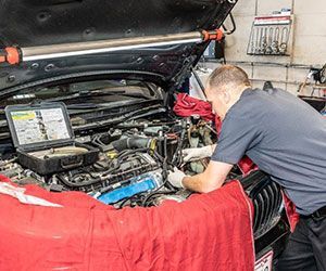 Mechanic working on a car engine in a garage; red cover, tools, overhead light.