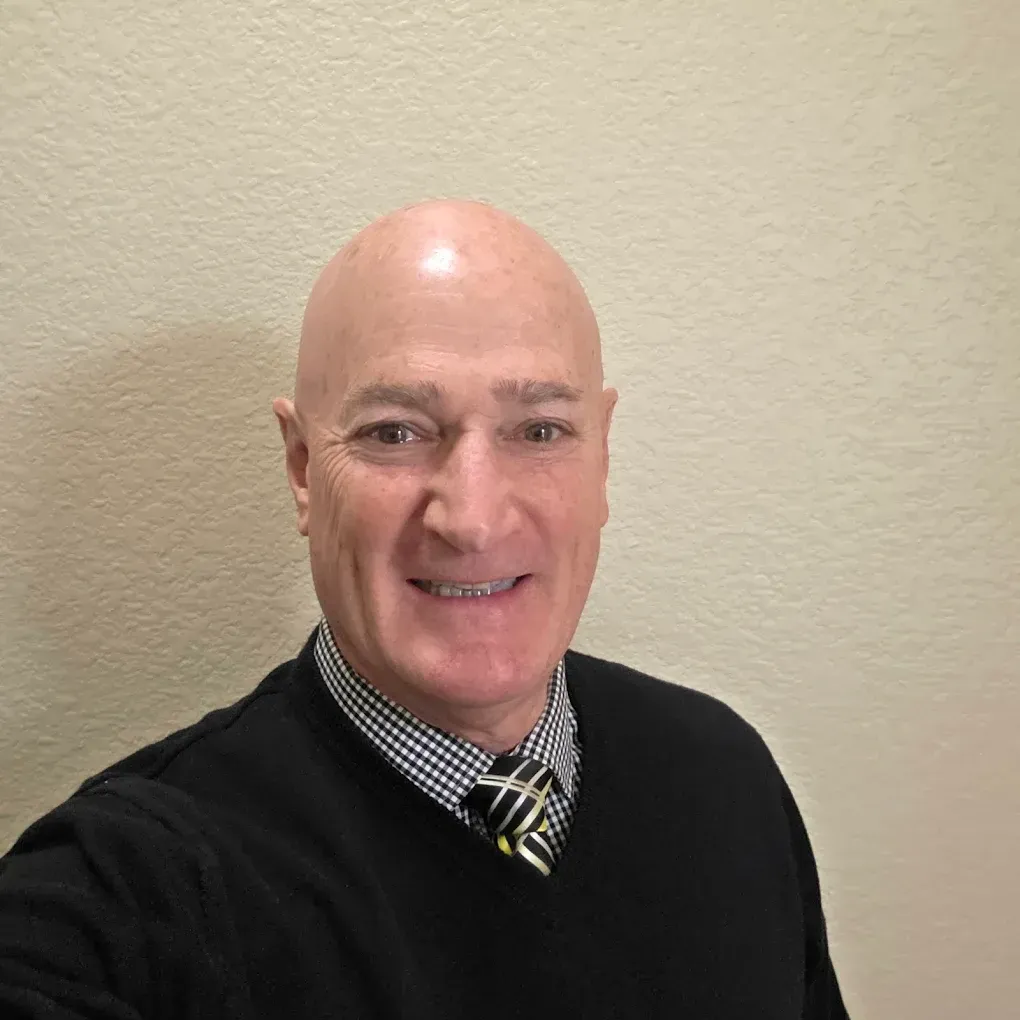 Smiling man in a black sweater and checkered shirt, standing against a beige wall.