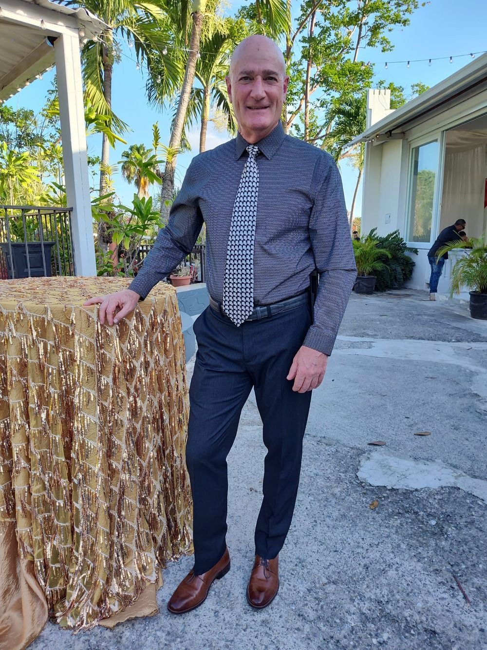 Man in a dark shirt and tie stands beside a gold-draped table outdoors in a sunny courtyard.