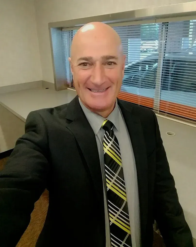 Person in a black suit and striped tie smiling in an office setting near windows.