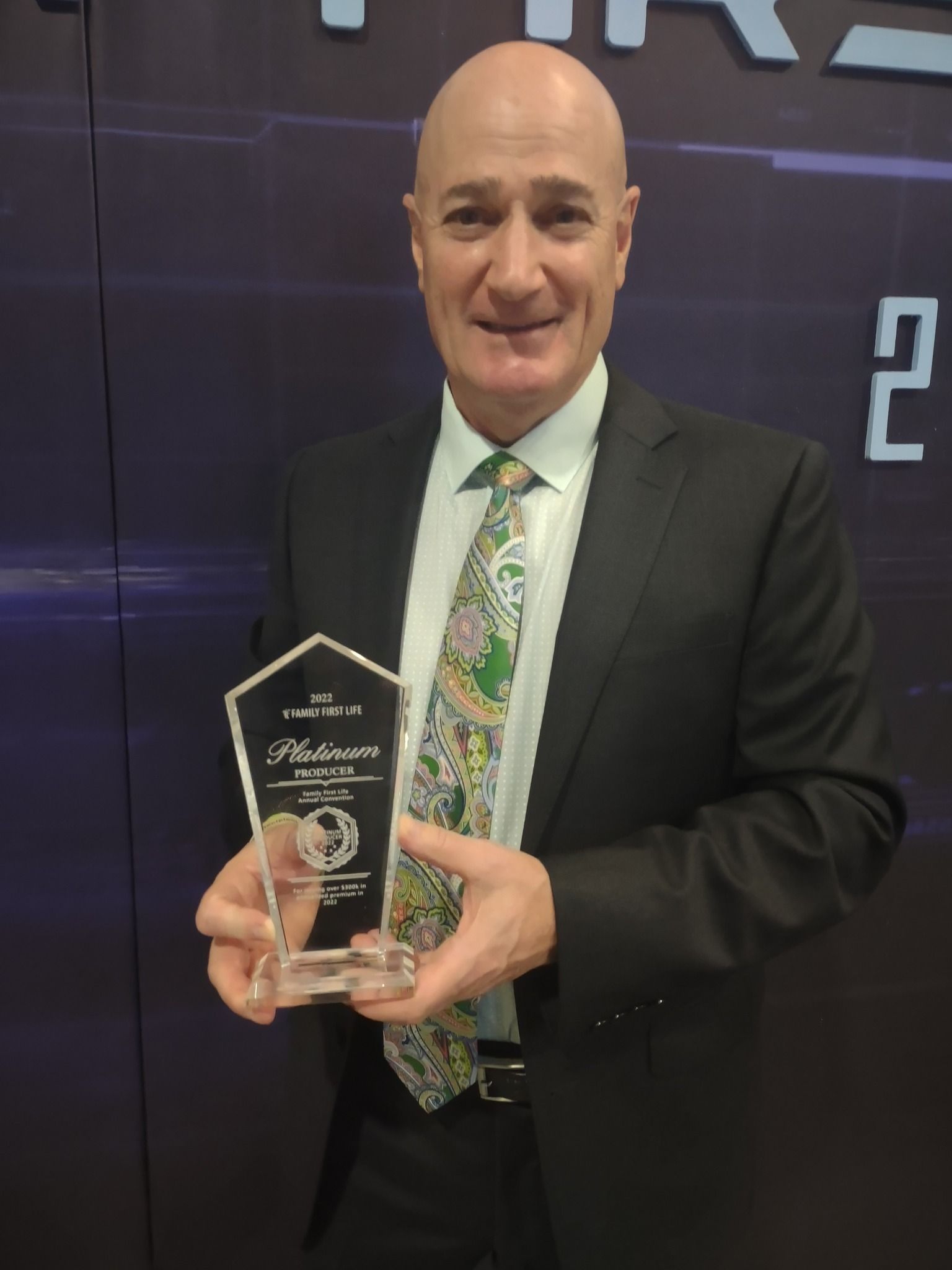 Man in a dark suit holding a clear award plaque with a green patterned tie against a dark backdrop