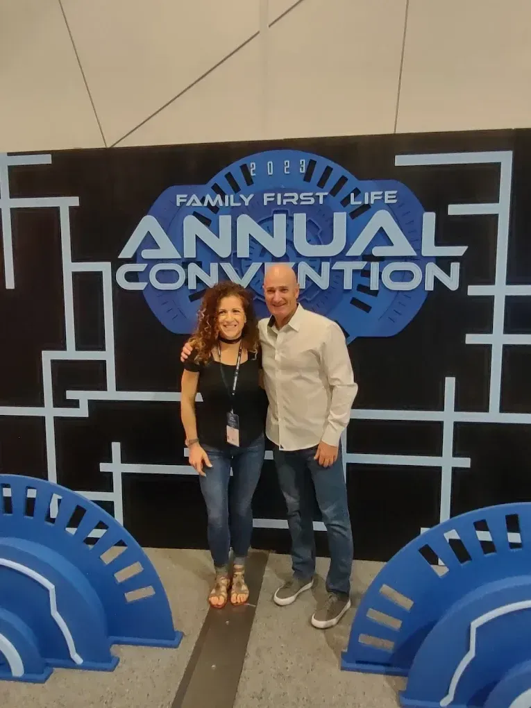Two people posing at a Family First Life Annual Convention backdrop with blue geometric decor.