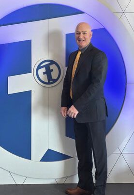 Man in a dark suit standing beside a large blue-and-white logo wall, smiling
