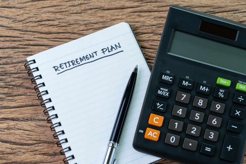Notebook labeled “Retirement Plan” beside a pen and calculator on a wooden desk
