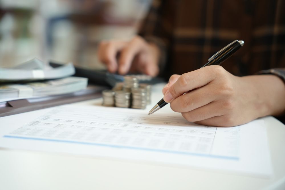 Hand signing a document beside a calculator and stacked papers on a desk