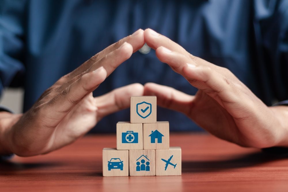 Hands forming a protective arch over stacked wooden blocks with icons for house, car, health, and travel safety.