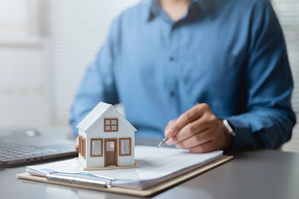 Person in blue shirt writing beside a small model house on a clipboard, suggesting home planning or real estate.