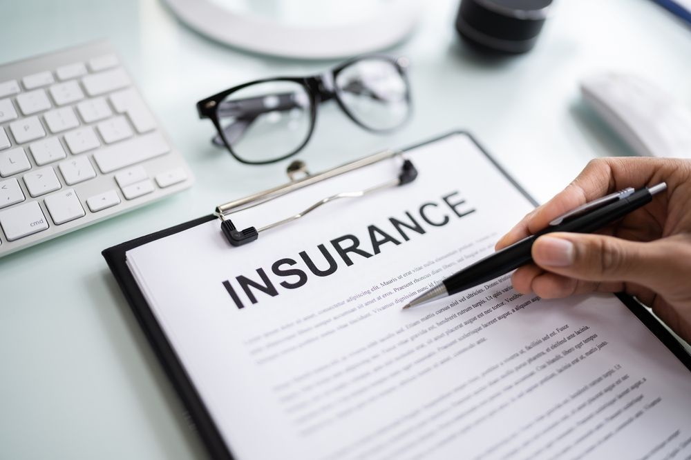Insurance document on clipboard being filled out by hand at a desk with keyboard and glasses nearby