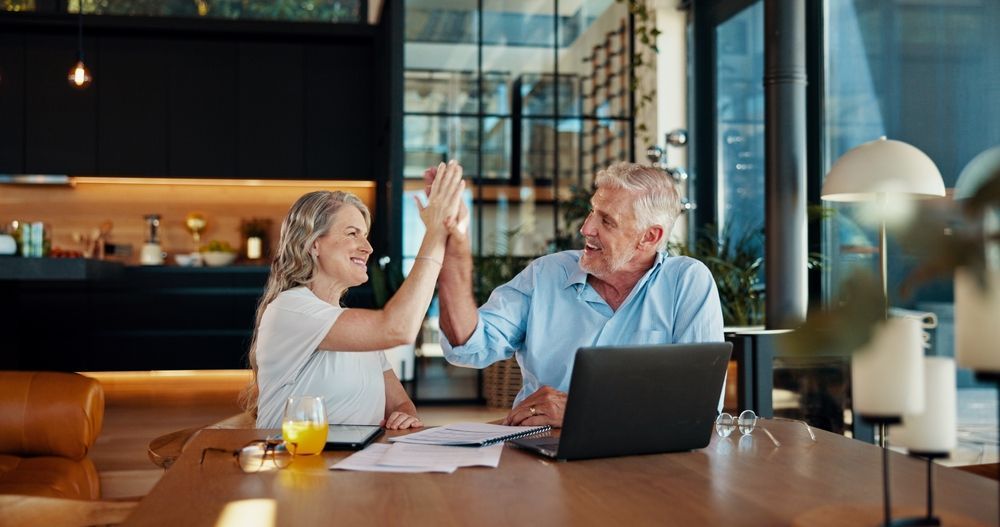 Two coworkers high-five at a café table with laptops and drinks, smiling in a bright office setting.