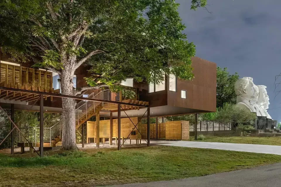 Modern house with wooden deck and large tree beside a grassy path at dusk.