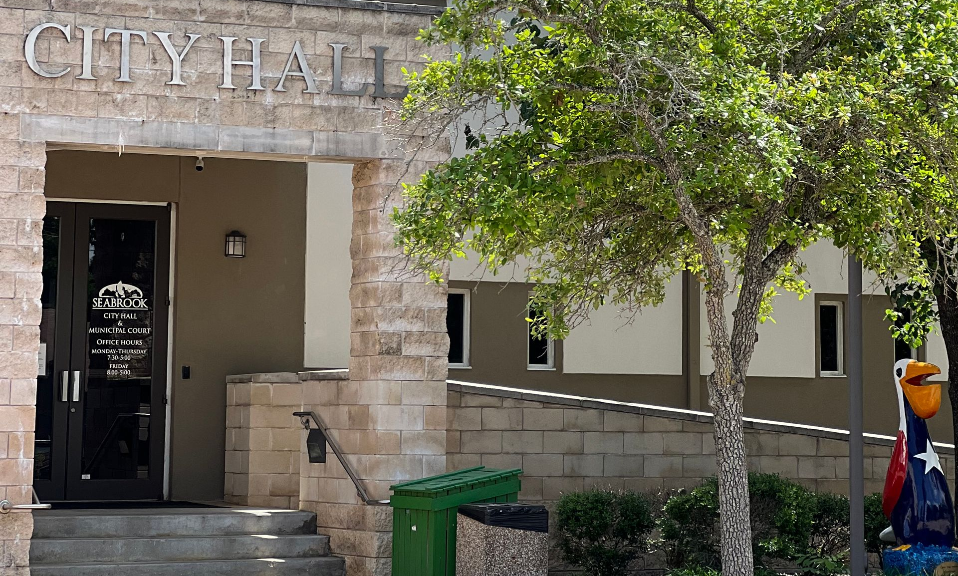 City hall entrance with stone facade, open doorway, tree, steps, and green trash bin