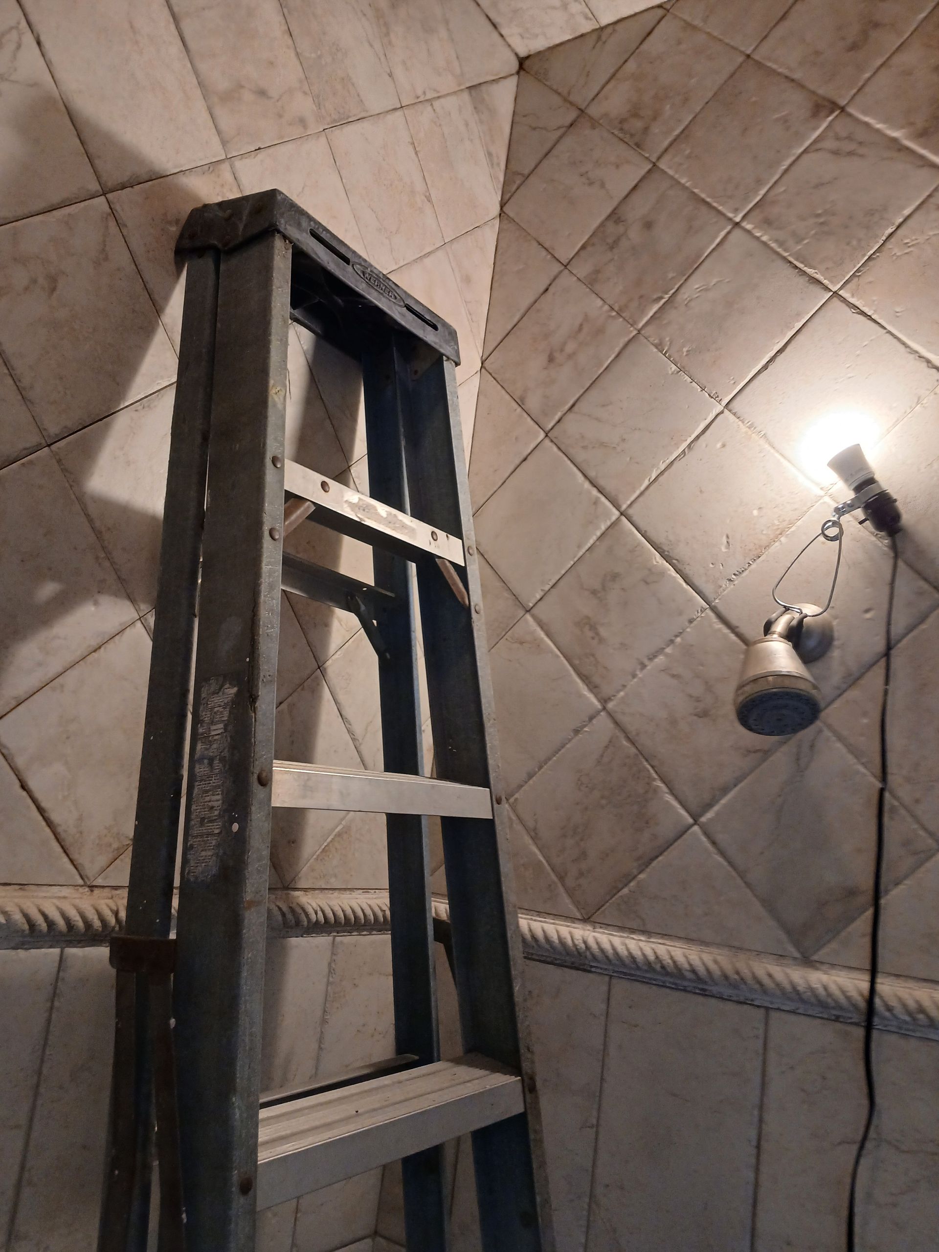 Ladder leaning against tiled wall, lit by a small spotlight, inside a room.