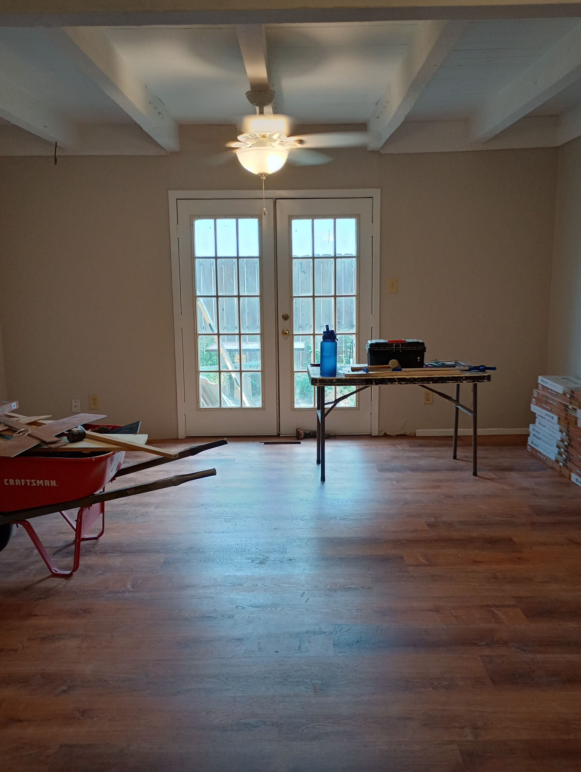Empty room with hardwood floors, French doors, ceiling fan, and a table with tools near the window.