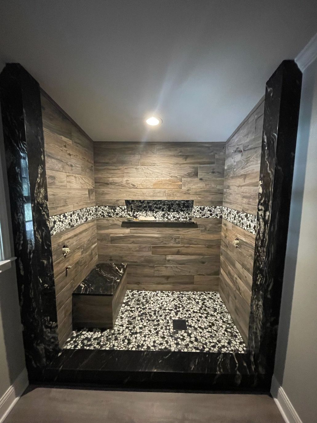Shower with wood-look tile walls, black and white mosaic floor, and dark stone pillars.