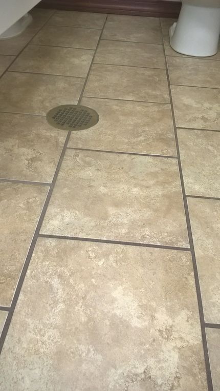 Bathroom floor with tan tiles, a drain, and a toilet in the corner.