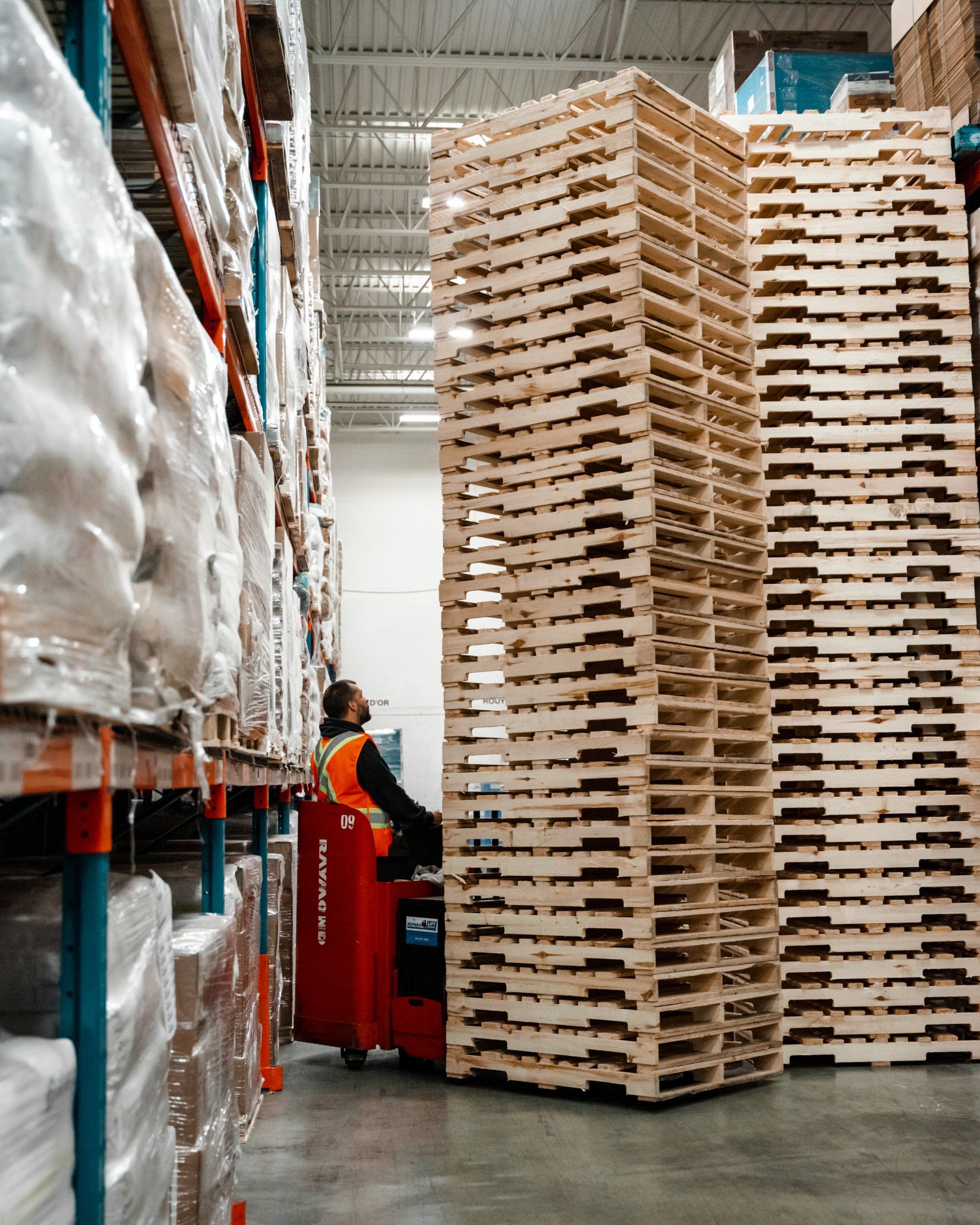 Un homme conduit un transpalette dans un entrepôt rempli de nombreuses palettes en bois.