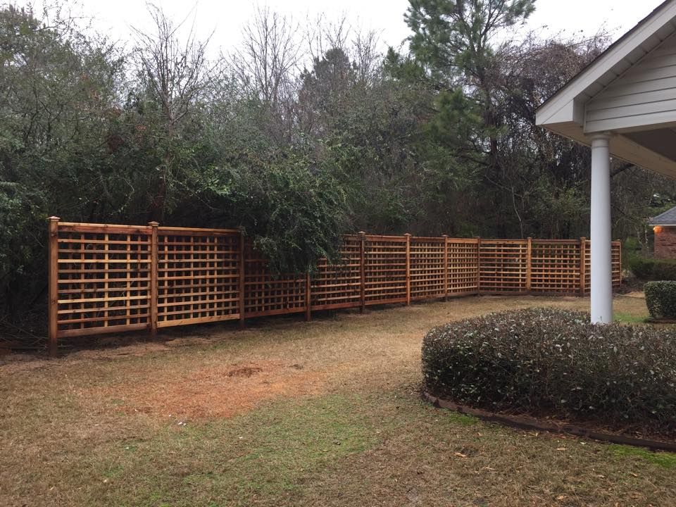 Wooden lattice fence curves along a yard, partially obscured by green bushes, adjacent to a house.