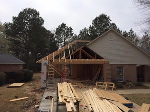 Construction of a garage addition on a house. Wooden framework visible, materials scattered around. Cloudy day.