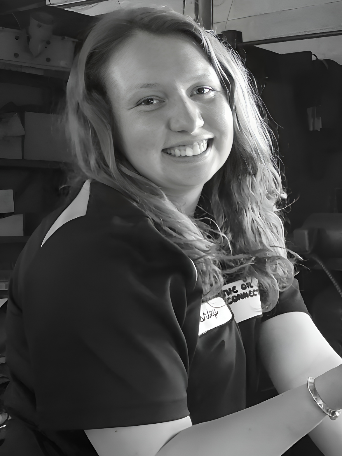 Woman with long hair smiling, wearing a dark shirt, with a bracelet, seated indoors.
