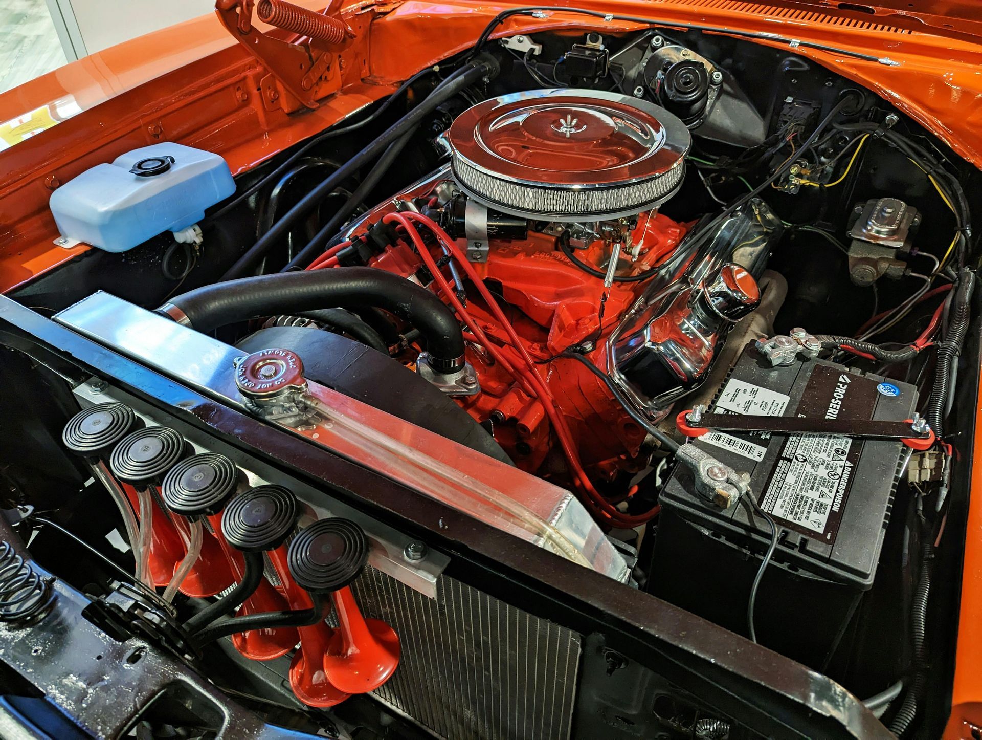 Orange muscle car engine, red intake, chrome air cleaner, black battery, and horns.